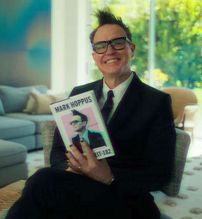 Mark Hoppus, seated in a living room, holds his Blink-182 book, smiling in a suit and glasses. Mark Hoppus, seated in a living room, holds his Blink-182 book, smiling in a suit and glasses.