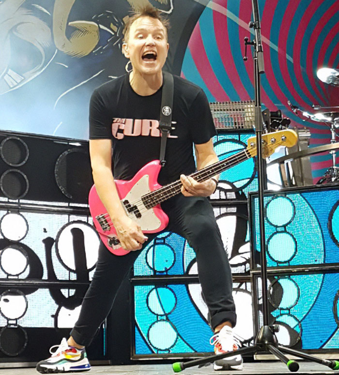 Mark Hoppus playing guitar on stage with a dynamic expression, wearing a "The Cure" shirt. Mark Hoppus playing guitar on stage with a dynamic expression, wearing a "The Cure" shirt.