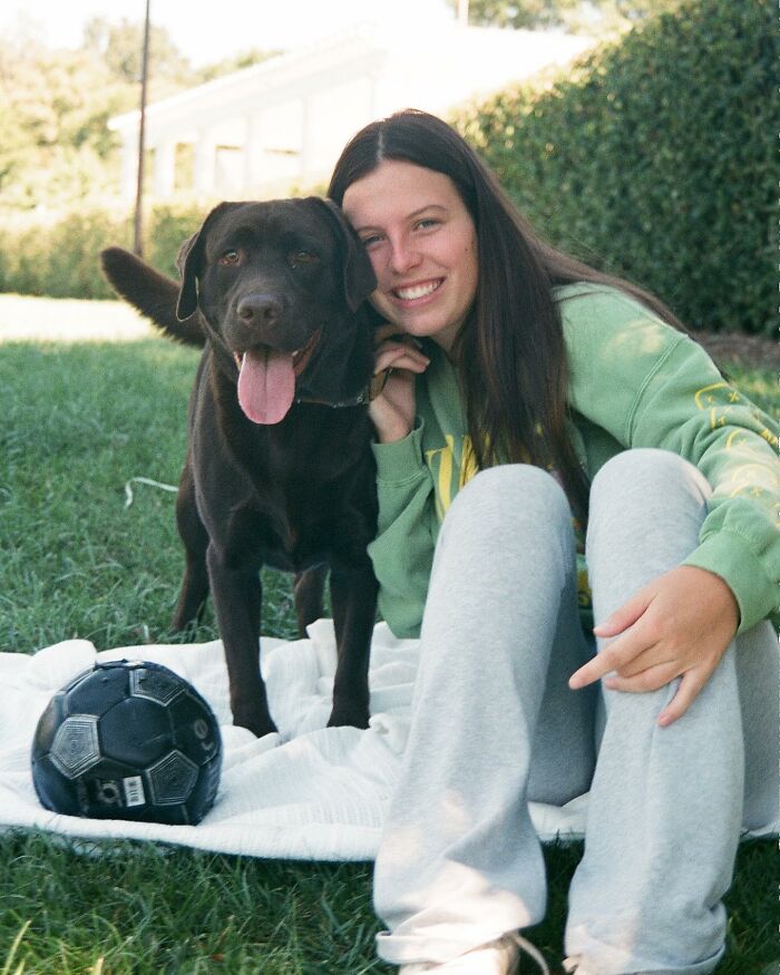 Young woman sitting on grass with Breuer the Labrador, smiling and posing beside a black soccer ball outdoors Young woman sitting on grass with Breuer the Labrador, smiling and posing beside a black soccer ball outdoors