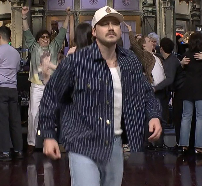 Man in a striped shirt and cap walking on stage during a lively SNL performance, surrounded by excited crowd reactions. Man in a striped shirt and cap walking on stage during a lively SNL performance, surrounded by excited crowd reactions.