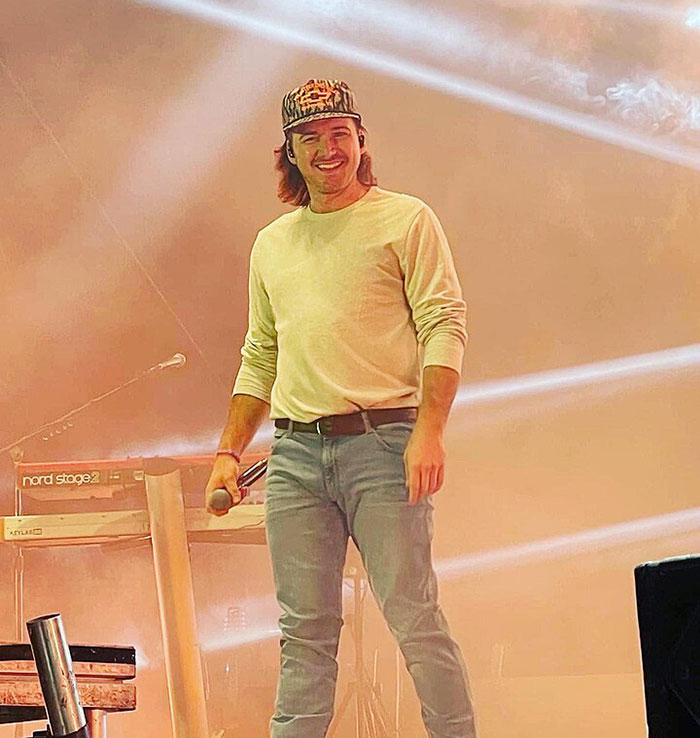 Man on stage holding microphone, wearing a casual outfit, with stage lights behind him. Man on stage holding microphone, wearing a casual outfit, with stage lights behind him.
