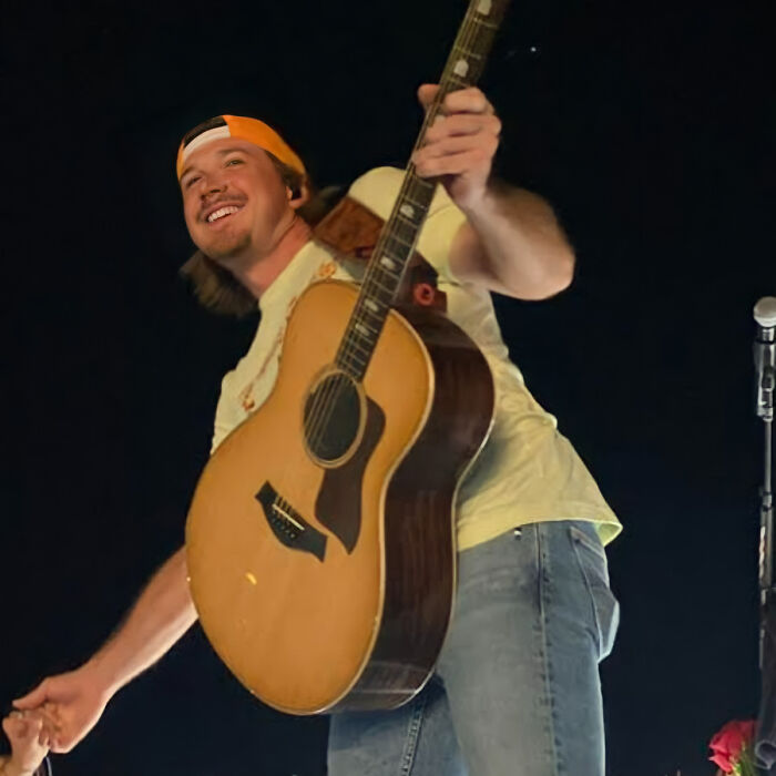 A smiling man in a backwards cap holds a guitar on stage, reaching out to the crowd. A smiling man in a backwards cap holds a guitar on stage, reaching out to the crowd.