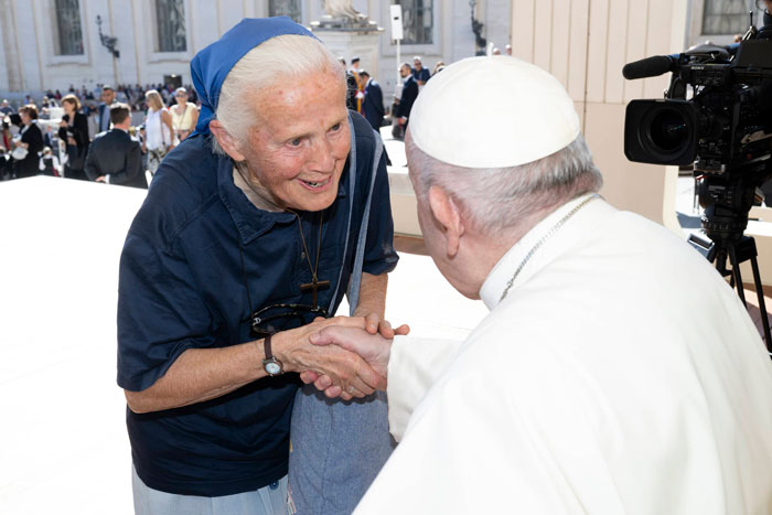 Nun smiling and shaking hands with the Pope, standing outdoors surrounded by people, camera capturing the moment. Nun smiling and shaking hands with the Pope, standing outdoors surrounded by people, camera capturing the moment.