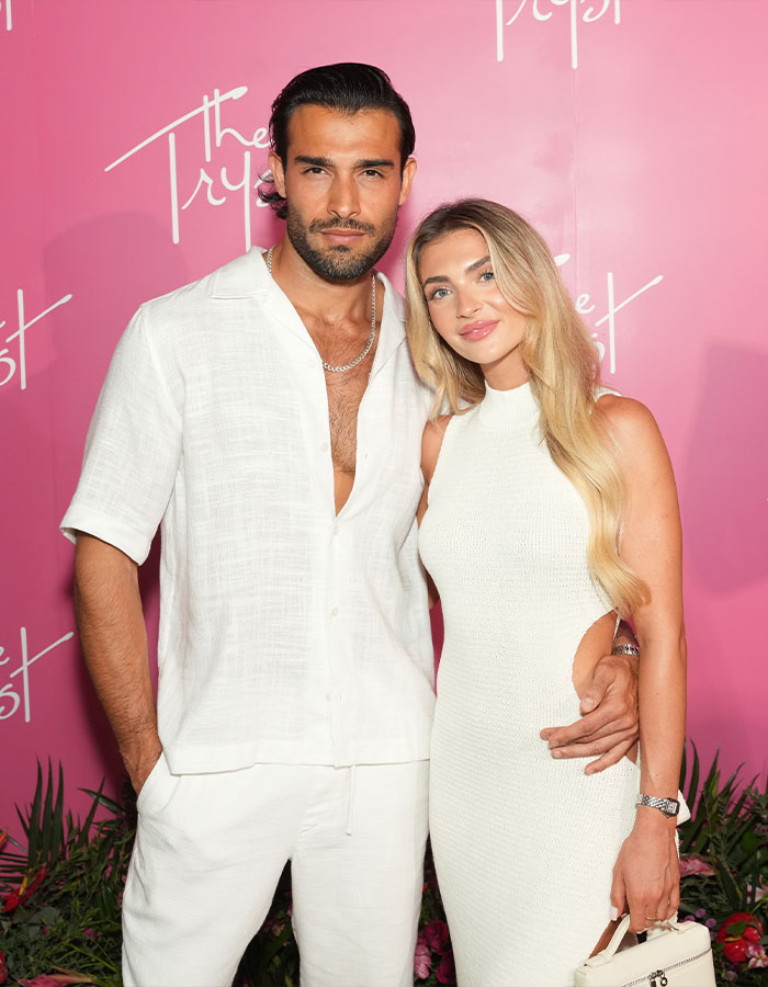 Man and woman in white attire at event, posing against pink backdrop. Man and woman in white attire at event, posing against pink backdrop.