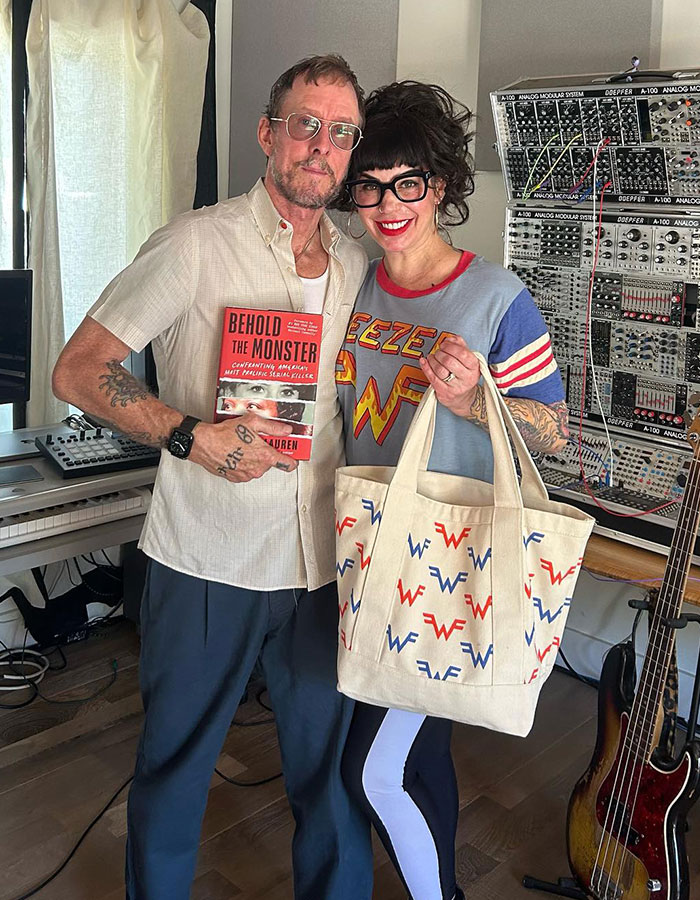 Weezer bassist in a studio, holding a book, next to a woman with a Weezer tote and shirt, near a music synthesizer. Weezer bassist in a studio, holding a book, next to a woman with a Weezer tote and shirt, near a music synthesizer.