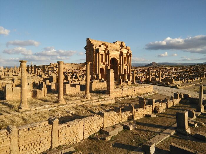 Ancient man-made structures with stone columns and ruins under a partly cloudy sky in a vast archaeological site.