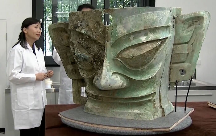 Scientist studies ancient artifact with a large, carved stone face in a lab setting.