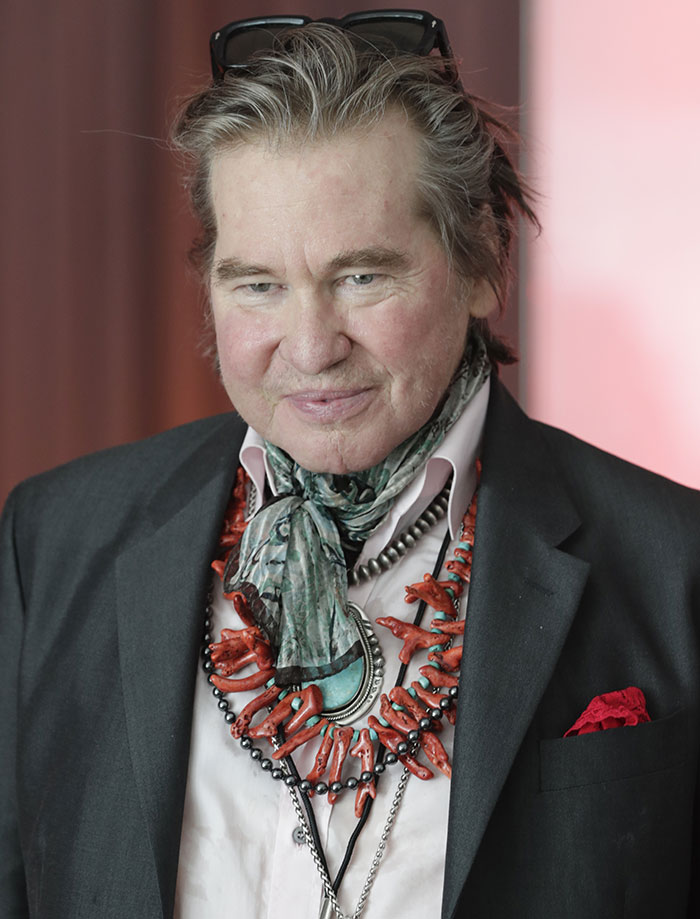 Man in a suit with a colorful necklace and scarf, wearing sunglasses on head and smiling gently in a formal setting. Man in a suit with a colorful necklace and scarf, wearing sunglasses on head and smiling gently in a formal setting.