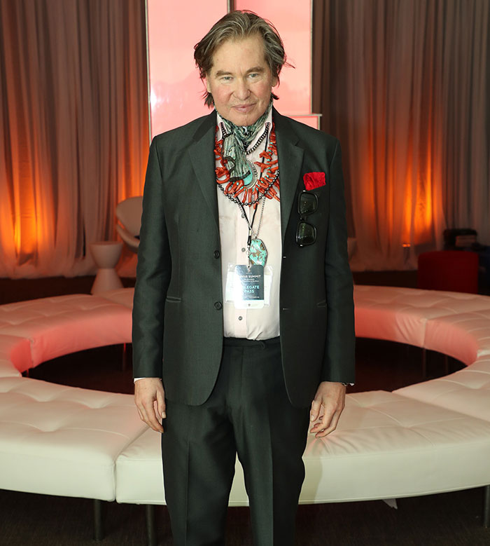 Legendary actor Val Kilmer in a suit, standing at an event with warm lighting in the background. Legendary actor Val Kilmer in a suit, standing at an event with warm lighting in the background.