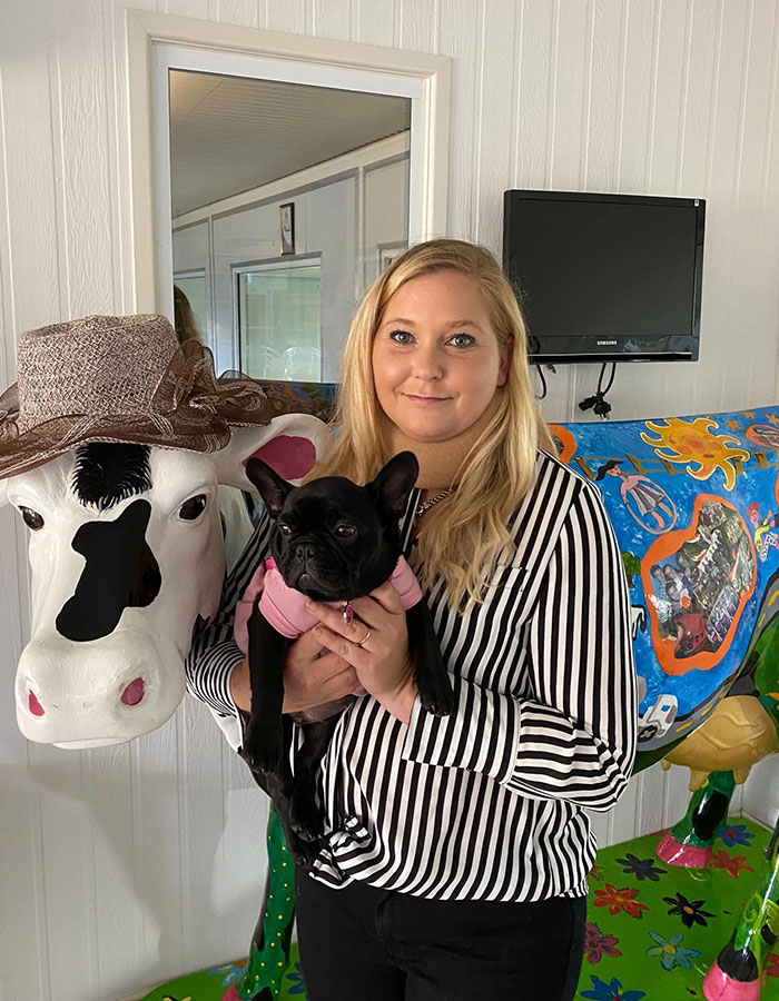Woman in striped shirt holding a black dog, standing next to a decorated cow sculpture. Woman in striped shirt holding a black dog, standing next to a decorated cow sculpture.