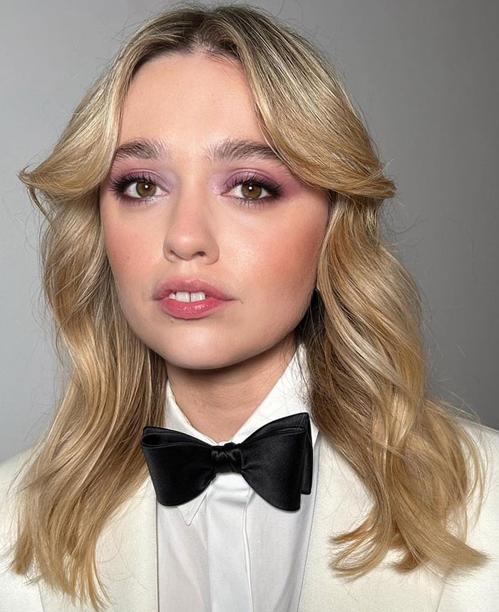 Blonde woman in a white outfit with a bow tie, associated with The White Lotus, highlighting her natural teeth. Blonde woman in a white outfit with a bow tie, associated with The White Lotus, highlighting her natural teeth.