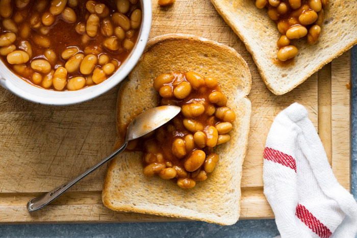 Beans on toast served with a side of baked beans and spoon, laid out on a wooden cutting board. Beans on toast served with a side of baked beans and spoon, laid out on a wooden cutting board.