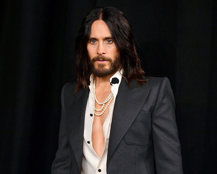 A person with long hair and beard, wearing a stylish suit and pearl necklace in front of a dark background. A person with long hair and beard, wearing a stylish suit and pearl necklace in front of a dark background.