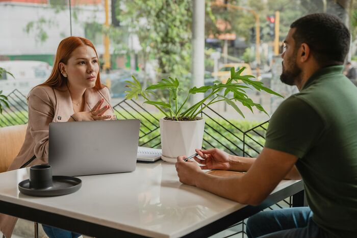 Two people discussing at a table with a laptop and plant, highlighting possible American tourist traits beyond accent.
