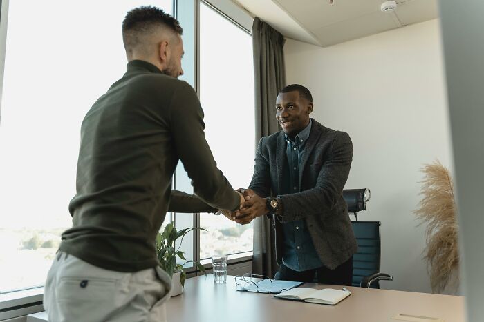 Two men shaking hands in an office setting, one seated and one standing, with business attire, indicating an American tourist.