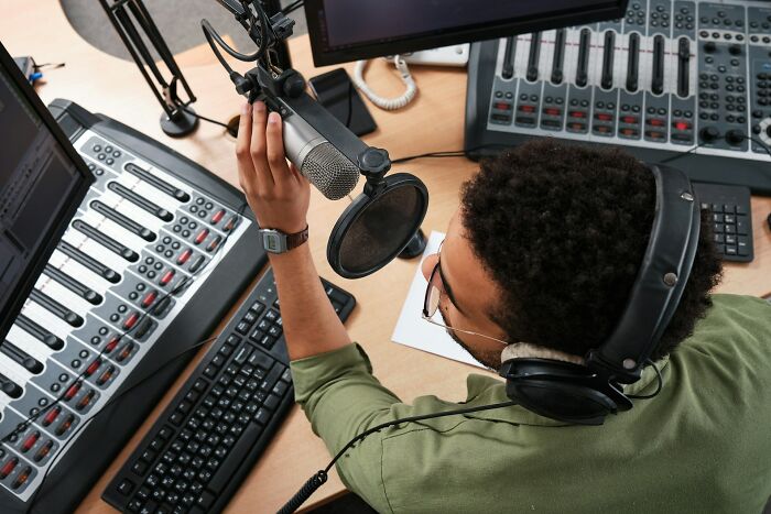 Radio host in studio wearing headphones and speaking into microphone, surrounded by audio equipment, preparing for broadcast.