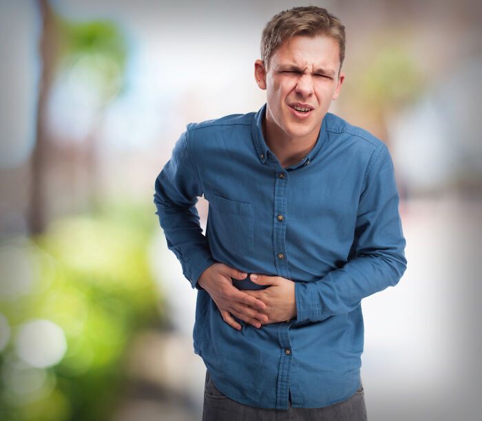 Young man enduring unexplainable pain, clutching stomach with a grimace while standing outdoors in a blurred setting