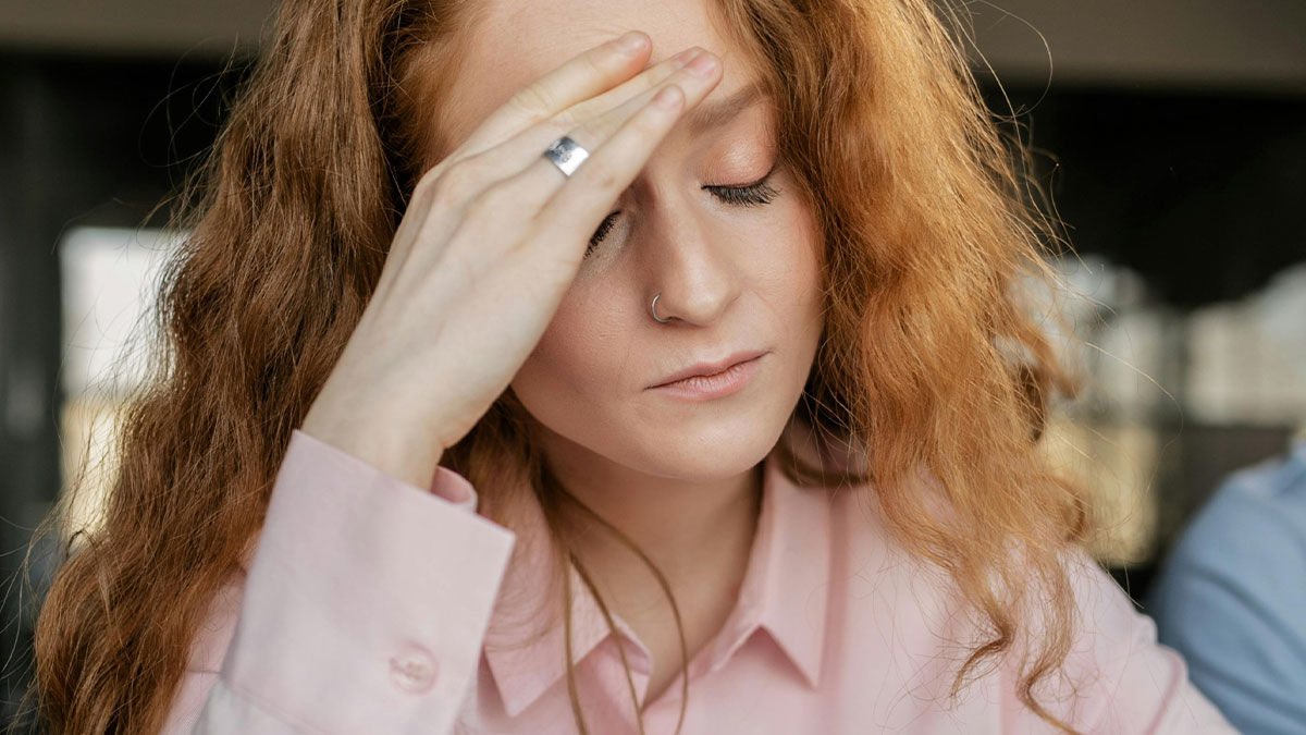 Woman with red hair and closed eyes, holding her forehead, appearing stressed with colleague blurred in background.