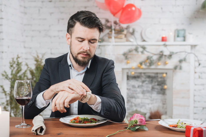 Man dressed in suit checking watch while sitting alone at dinner table with wine glass and rose nearby. Man dressed in suit checking watch while sitting alone at dinner table with wine glass and rose nearby.