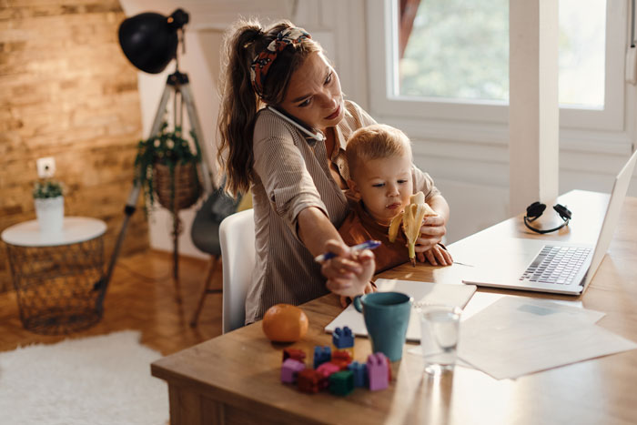Woman babysitting sister’s kids, working on laptop at dining table while toddler eats a banana and plays with toys.