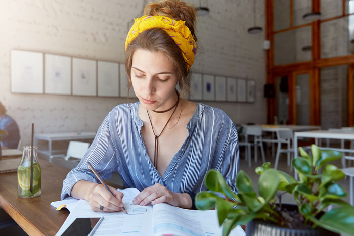 Young woman babysitting sister’s kids while working on paperwork at a table with plants and a drink nearby.