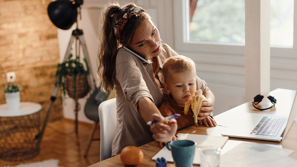 Woman multitasking babysitting her sister's kid while working from home at a desk with laptop and phone.