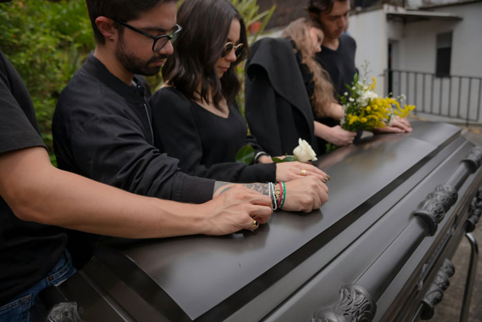 Mourners in black clothes standing around a casket, holding flowers, showing grief at a somber funeral gathering. Mourners in black clothes standing around a casket, holding flowers, showing grief at a somber funeral gathering.