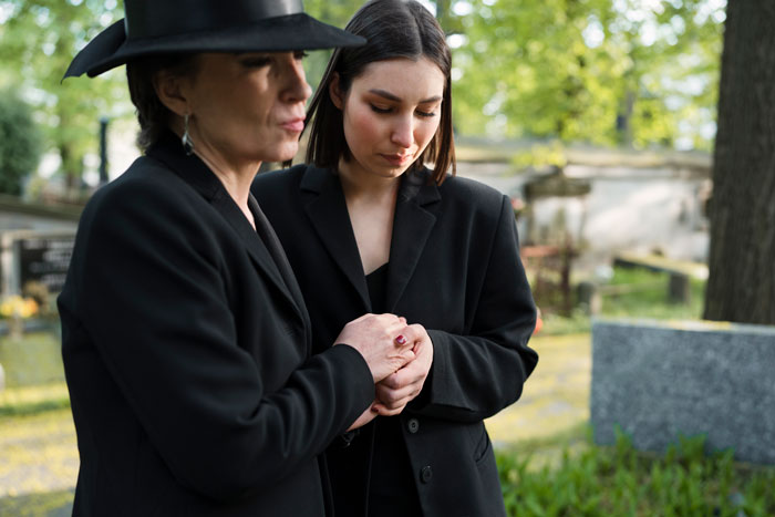 Two women in black, one comforting the other at a cemetery, amid a somber setting. Two women in black, one comforting the other at a cemetery, amid a somber setting.
