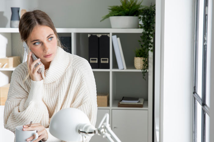 Woman in white sweater looking concerned while talking on phone, holding a mug, near a window in a cozy room. Woman in white sweater looking concerned while talking on phone, holding a mug, near a window in a cozy room.