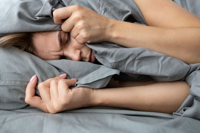 Woman struggling with lack of proper sleep, covering face with pillow, showing signs of stress and exhaustion in bed.