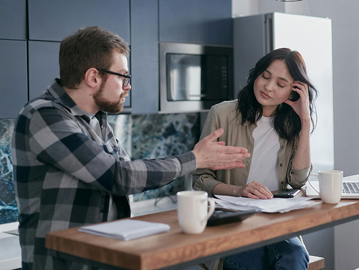 Man discussing at a kitchen table, woman with hand on face, appearing frustrated about medical expertise challenge. Man discussing at a kitchen table, woman with hand on face, appearing frustrated about medical expertise challenge.