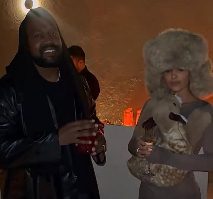 Party scene with Bianca Censori in a furry hat holding a drink, next to a man in a dark outfit, in a dimly lit setting. Party scene with Bianca Censori in a furry hat holding a drink, next to a man in a dark outfit, in a dimly lit setting.