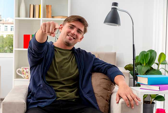 Young man in a cozy room points playfully at the camera, sitting on a couch near a lamp and bookshelf. Young man in a cozy room points playfully at the camera, sitting on a couch near a lamp and bookshelf.