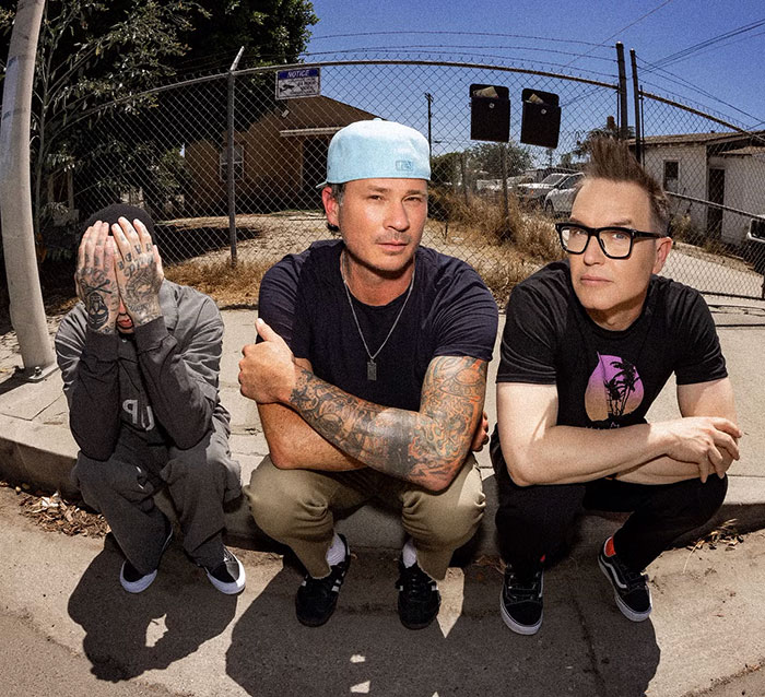 Blink-182's Mark Hoppus sits on curb with bandmates in casual attire, outdoors near a fence. Blink-182's Mark Hoppus sits on curb with bandmates in casual attire, outdoors near a fence.