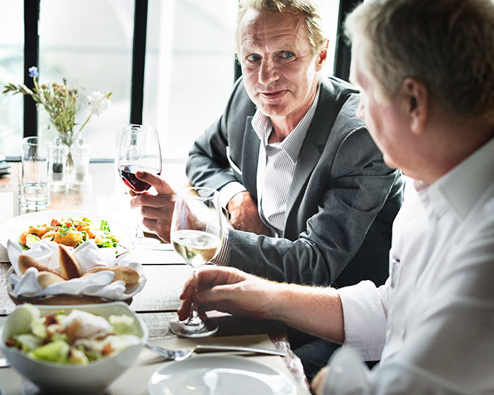 Two men dining, one in a suit, holding wine glasses, discussing traders’ arrogance in a restaurant setting. Two men dining, one in a suit, holding wine glasses, discussing traders’ arrogance in a restaurant setting.