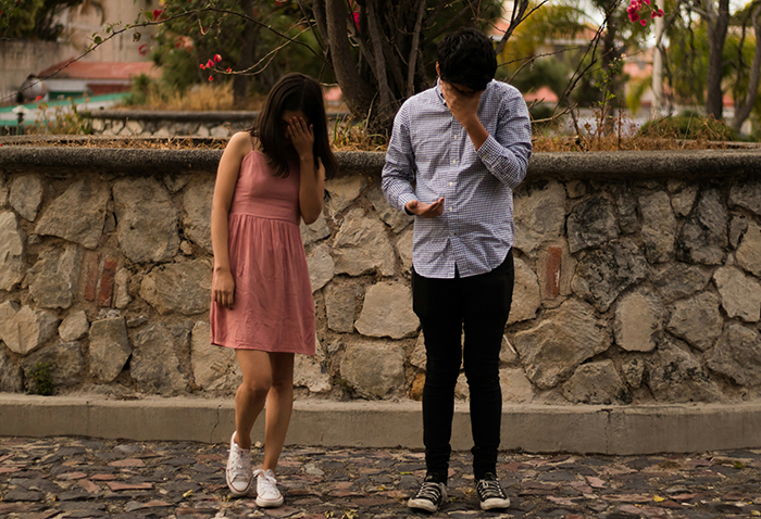 A woman and man stand apart, both covering their faces, in a park setting with stone wall and flowers. A woman and man stand apart, both covering their faces, in a park setting with stone wall and flowers.