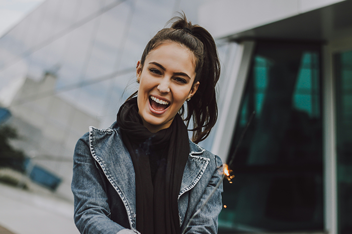 Woman smiling brightly in front of a modern building, wearing a denim jacket and black scarf. Woman smiling brightly in front of a modern building, wearing a denim jacket and black scarf.