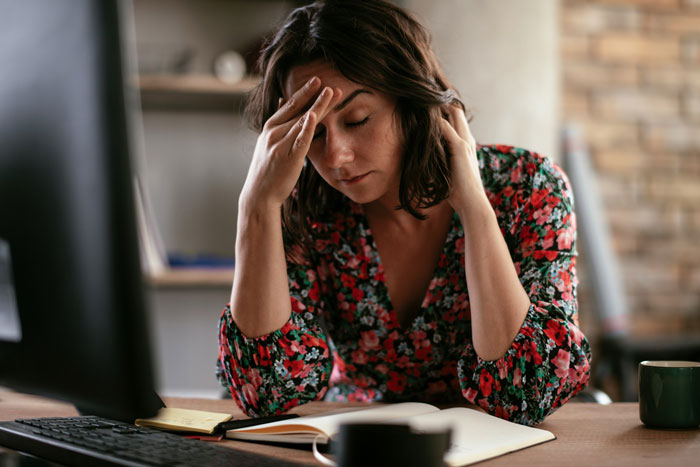 A stressed woman in a floral dress holds her head in frustration at a desk. A stressed woman in a floral dress holds her head in frustration at a desk.