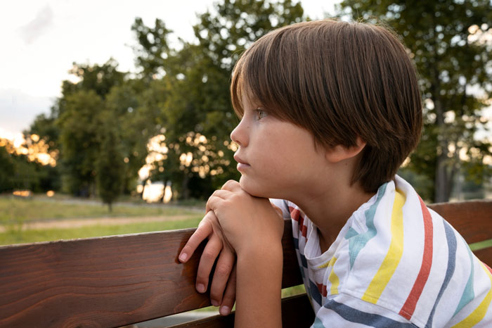 Young boy in a striped shirt sitting on a bench outdoors, reflecting on a woman buying a swing set for her kid. Young boy in a striped shirt sitting on a bench outdoors, reflecting on a woman buying a swing set for her kid.
