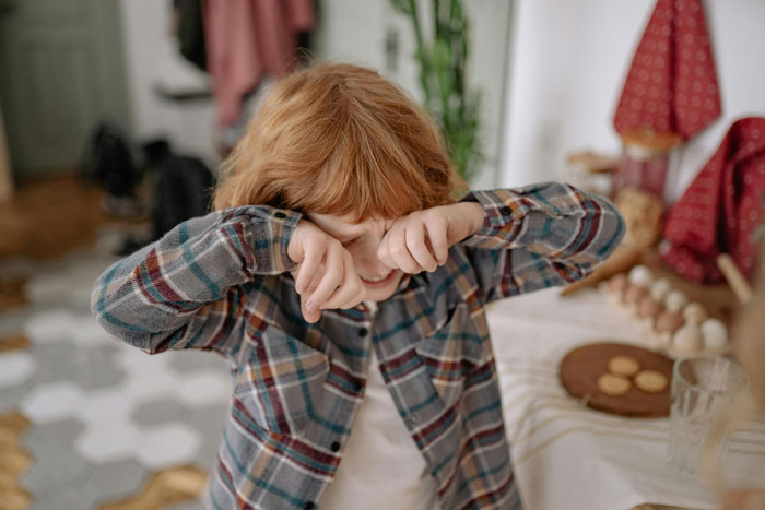 Young red-haired child in plaid shirt wiping eyes indoors, illustrating impact of wealth flexing on childhood experiences. Young red-haired child in plaid shirt wiping eyes indoors, illustrating impact of wealth flexing on childhood experiences.
