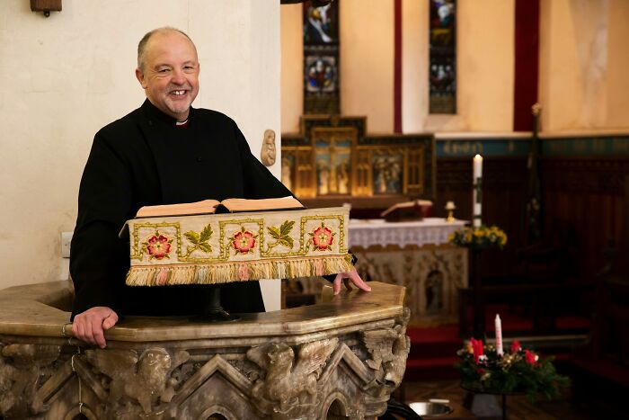 Priest smiling at a church pulpit, wearing a black robe, surrounded by ornate decor and candles.