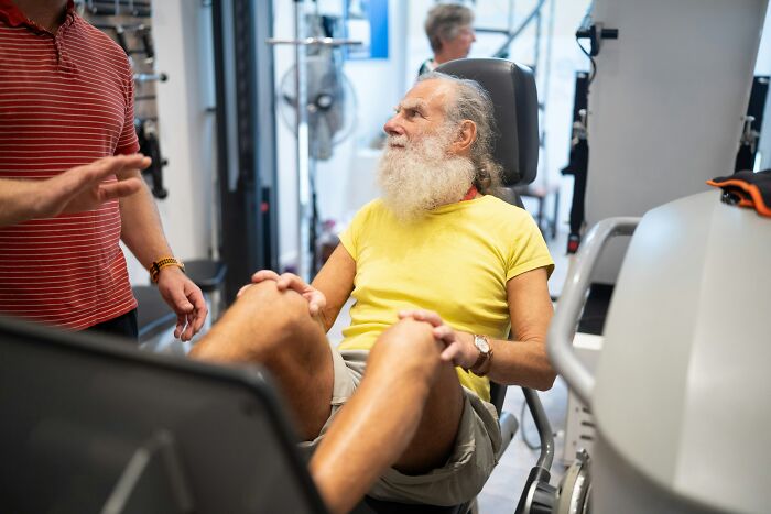 Bearded man in yellow shirt sitting on a gym machine, engaging with another person, possibly discussing adult tantrums.
