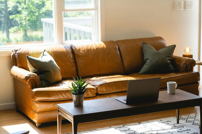 Cozy living room with a brown leather couch, green pillows, and a laptop on a wooden coffee table in sunlight.
