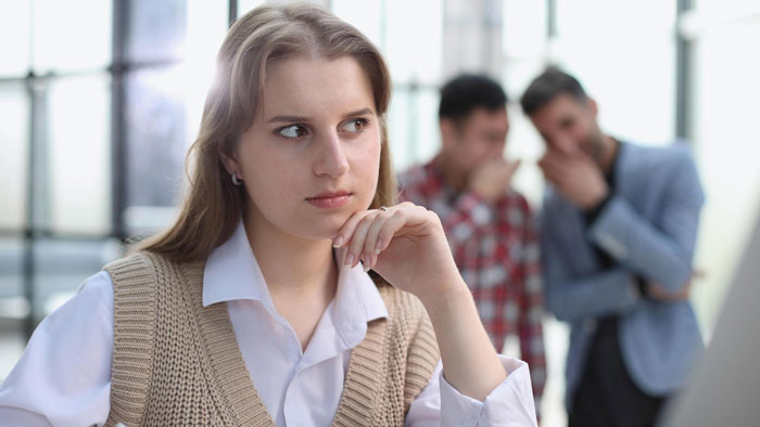 Woman in an office setting looks concerned while two coworkers talk in the background during a video call. Woman in an office setting looks concerned while two coworkers talk in the background during a video call.
