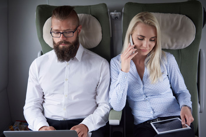 Couple seated in first class, the man using a laptop and the woman on the phone with a tablet, appearing entitled. Couple seated in first class, the man using a laptop and the woman on the phone with a tablet, appearing entitled.