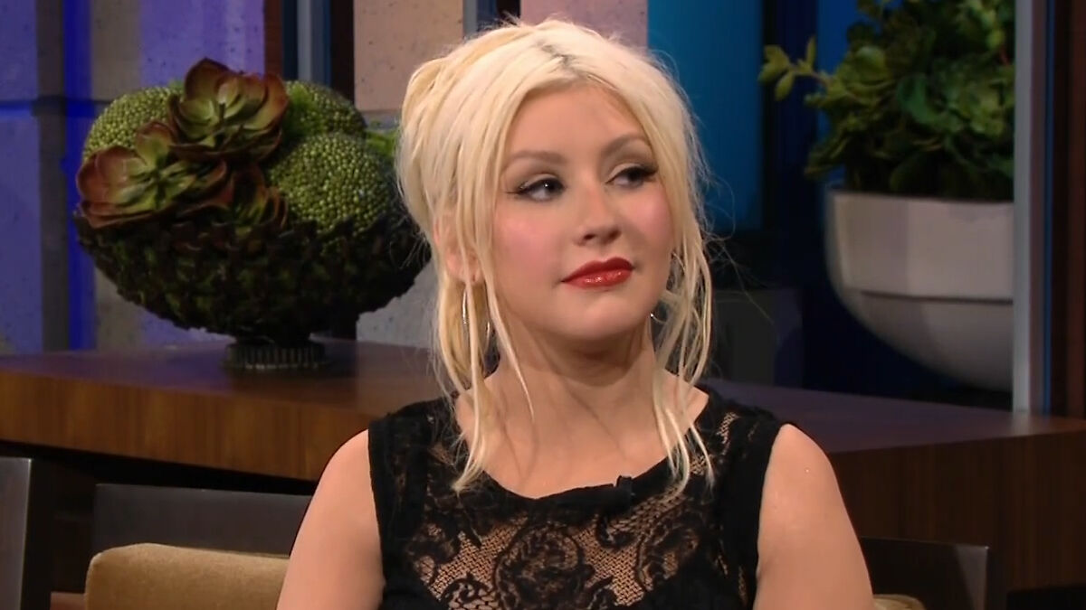 Christina Aguilera wearing a black lace dress, with styled blonde hair, in a TV interview setting.