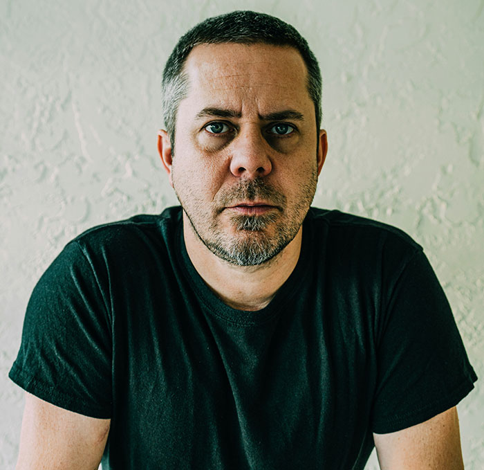 Man in a black shirt with a serious expression, in front of a textured wall, illustrating a client's delusional state. Man in a black shirt with a serious expression, in front of a textured wall, illustrating a client's delusional state.