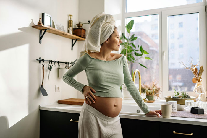 Pregnant woman in a kitchen, hand on hip, wearing a towel on her head, near a window and plants. Pregnant woman in a kitchen, hand on hip, wearing a towel on her head, near a window and plants.