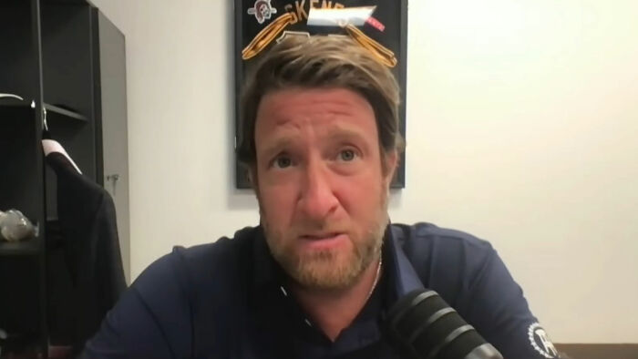 A person in a blue shirt speaking into a microphone, framed sports memorabilia in the background. A person in a blue shirt speaking into a microphone, framed sports memorabilia in the background.