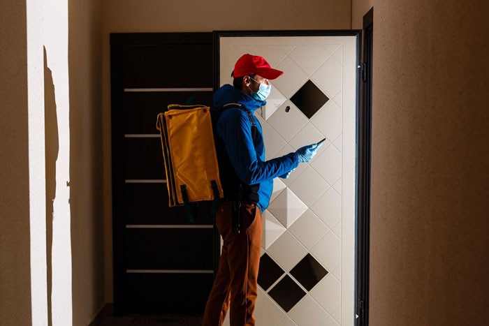 Delivery company driver with a yellow bag at a white door, wearing a red cap and mask, in a dimly lit hallway. Delivery company driver with a yellow bag at a white door, wearing a red cap and mask, in a dimly lit hallway.
